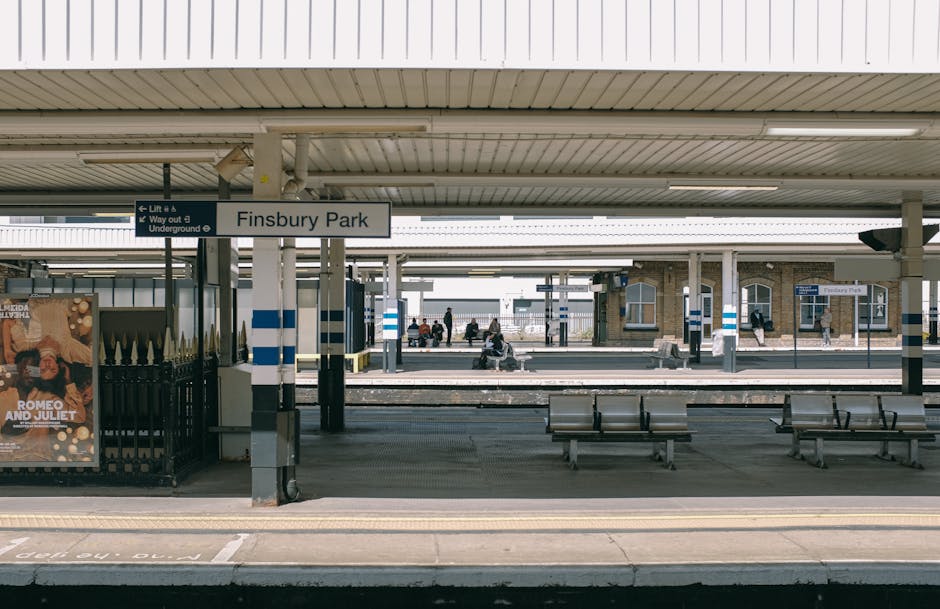 A view of Finsbury Park railway station platform showing seating areas with metal benches, a metal fence on the left side, and a large sign above indicating the station name as 'Finsbury Park.' The platform is covered by a roof with fluorescent lighting, and in the background, there are several people sitting or walking along the platform. The station features a clean, tiled environment with a mix of brick and metal structures, and the perspective suggests a typical setting for home relocation and furniture transport services provided by Bromley Movers, with the station serving as a key point for moving logistics and local moves in the Bromley area.