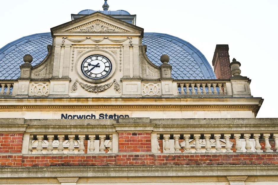 A close-up view of Norwich Station's clock tower and ornate architectural façade, constructed from light-colored stone, with decorative elements such as carved garlands and ornamental balls on the roofline, set against an overcast sky. Below the clock, the station's name is displayed on a white panel, with the background showcasing a red brick wall. On the right side, part of a chimney with a brick finish and a chimney pot is visible. The scene captures a part of the building's upper exterior, which is involved in a home relocation process by Bromley Movers, featuring moving blankets, cardboard boxes, and a trolley visible on the pavement below, ready for furniture transport and loading onto a van for the next phase of the packing and moving service.