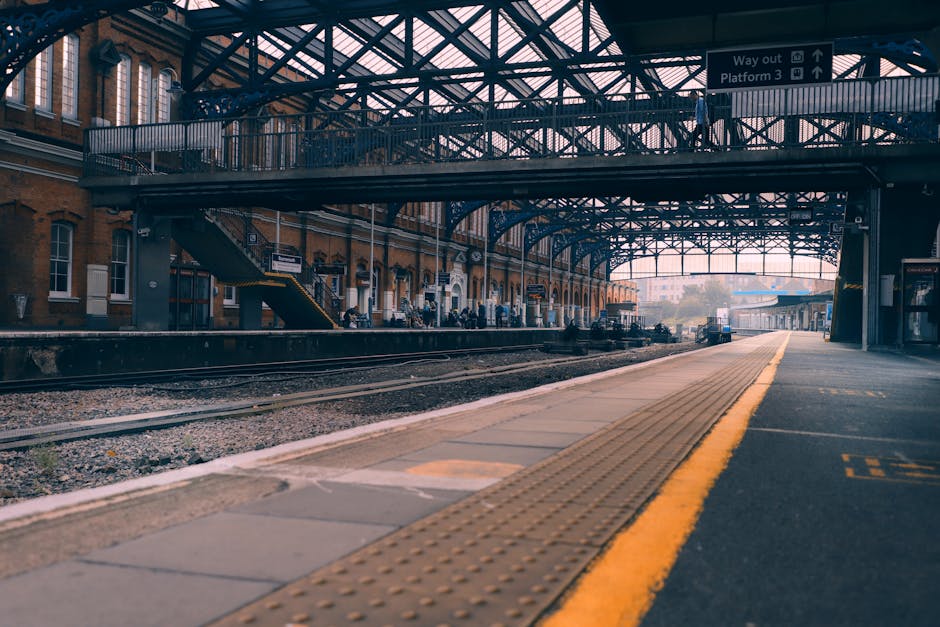 A view of Finsbury Park railway station platform showing seating areas with metal benches, a metal fence on the left side, and a large sign above indicating the station name as 'Finsbury Park.' The platform is covered by a roof with fluorescent lighting, and in the background, there are several people sitting or walking along the platform. The station features a clean, tiled environment with a mix of brick and metal structures, and the perspective suggests a typical setting for home relocation and furniture transport services provided by Bromley Movers, with the station serving as a key point for moving logistics and local moves in the Bromley area.