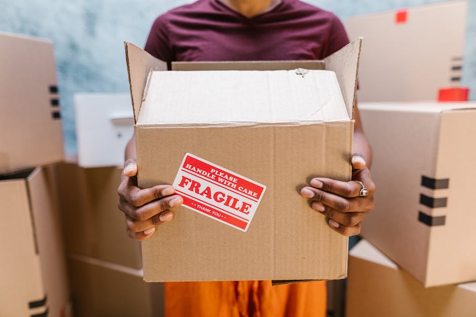 A young woman with long brown hair smiling while holding an open cardboard box labeled 'FRAGILE GLASS' during a home relocation process. She is standing indoors against a textured blue wall, surrounde