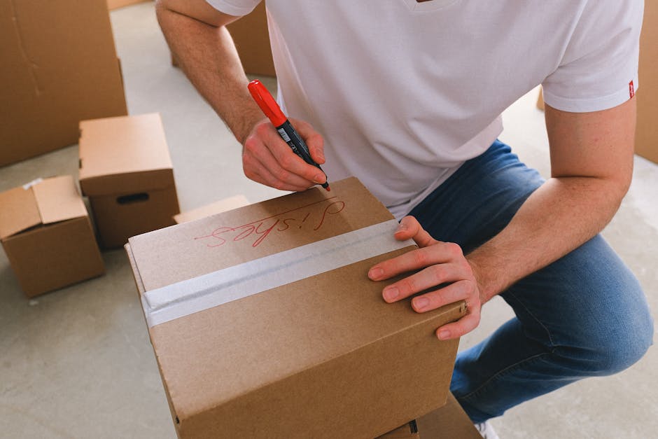 A person dressed in a white t-shirt and blue jeans is kneeling on the floor, writing the word 'Stairs' in red marker on a rectangular cardboard box that is sealed with white packing tape. The box is situated among several other similarly sealed cardboard boxes and is part of a home relocation process. The scene takes place inside a room with a light-colored floor, and a doorway with an open door is visible in the background. The person appears to be preparing the box for careful handling or labeling as part of professional removals services, such as those provided by Bromley Movers, specializing in packing and moving for house relocations. The environment suggests an organized packing setup, with supplies and boxing materials ready for transport, and the image captures the careful flow of logistics involved in furniture transport and moving preparations.