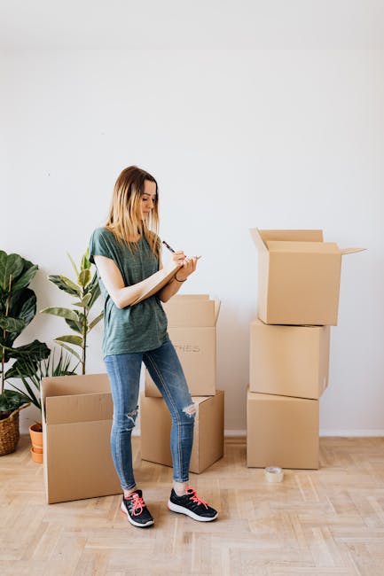 A young woman with shoulder-length blonde hair, wearing a casual green t-shirt, ripped blue jeans, and black and pink sneakers, stands inside a bright room with white walls and wooden flooring. She is holding a notepad and pen, appearing to take notes during a home relocation planning process. Behind her, there are several cardboard moving boxes of various sizes, some stacked with open lids, ready for packing belongings. To her left, a tall potted plant with broad green leaves is positioned on the floor, adding a natural element to the scene. The setting suggests involvement in busy packing and moving activities, typical of house removals and furniture transport, with the boxes prepared for transportation or storage. The image is relevant to the services offered by Bromley Movers, matching the context of packing and removals for a property on Bromley High Street, as referenced in the webpage about packing and removals checklist.